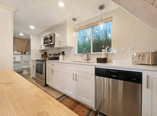 Modern kitchen with white cabinets, stainless steel appliances, and a window overlooking greenery. Cozy and well-lit space.